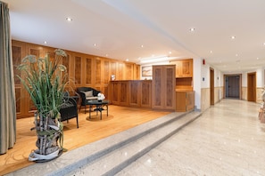 Um saguão de hotel com painéis de madeira, um balcão de recepção, uma área de estar com uma cadeira e mesa, e um vaso de planta.