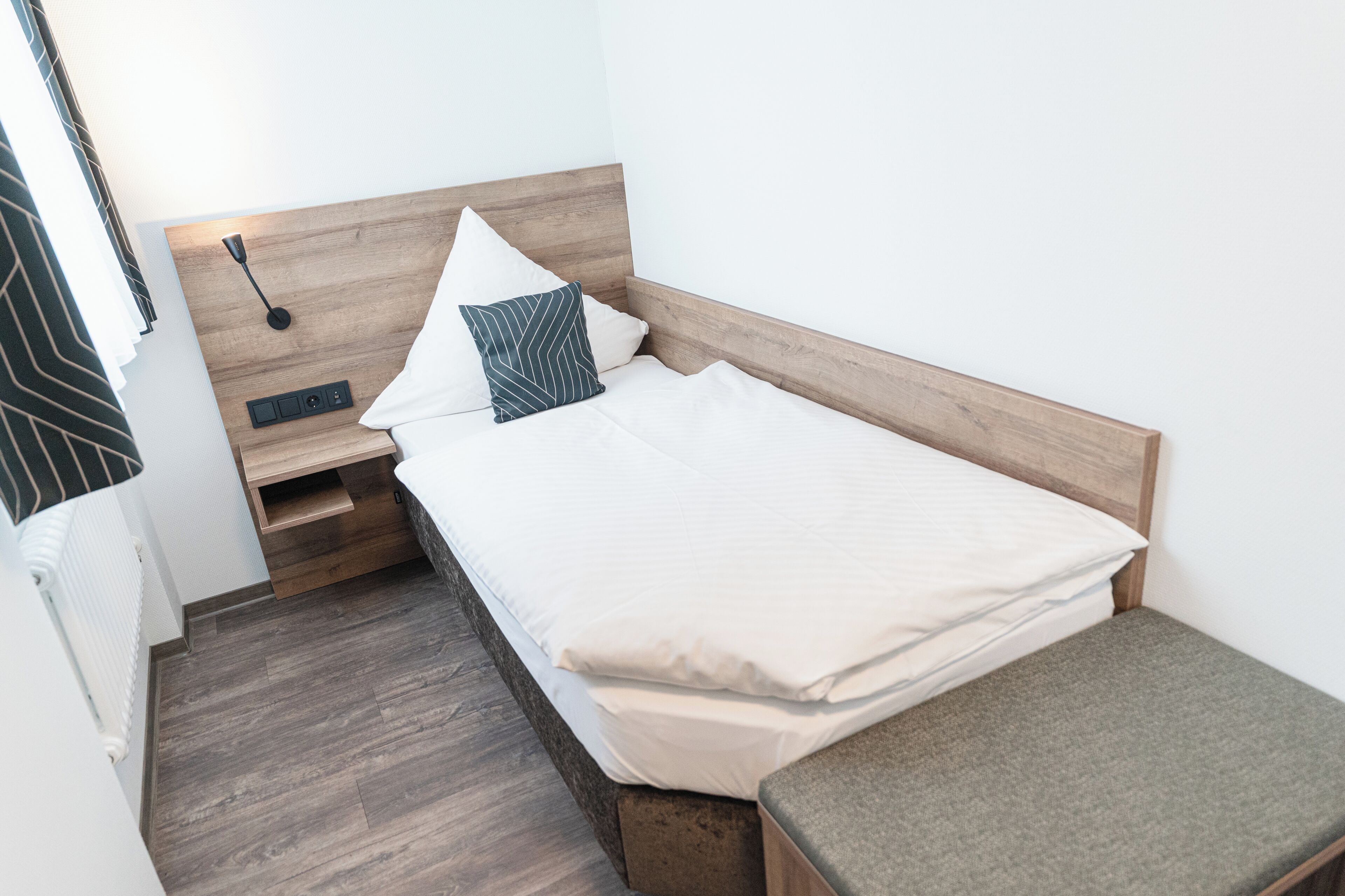 Una cama pequeña y ordenada, con cabecera de madera, una mesita de noche y un banco.