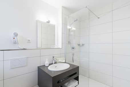 Ein modernes Badezimmer mit einem weißen Waschbecken, einem dunklen Waschtisch, einem großen Spiegel und einer Duschkabine aus Glas.. Hotel Gasthof Zum Rössle