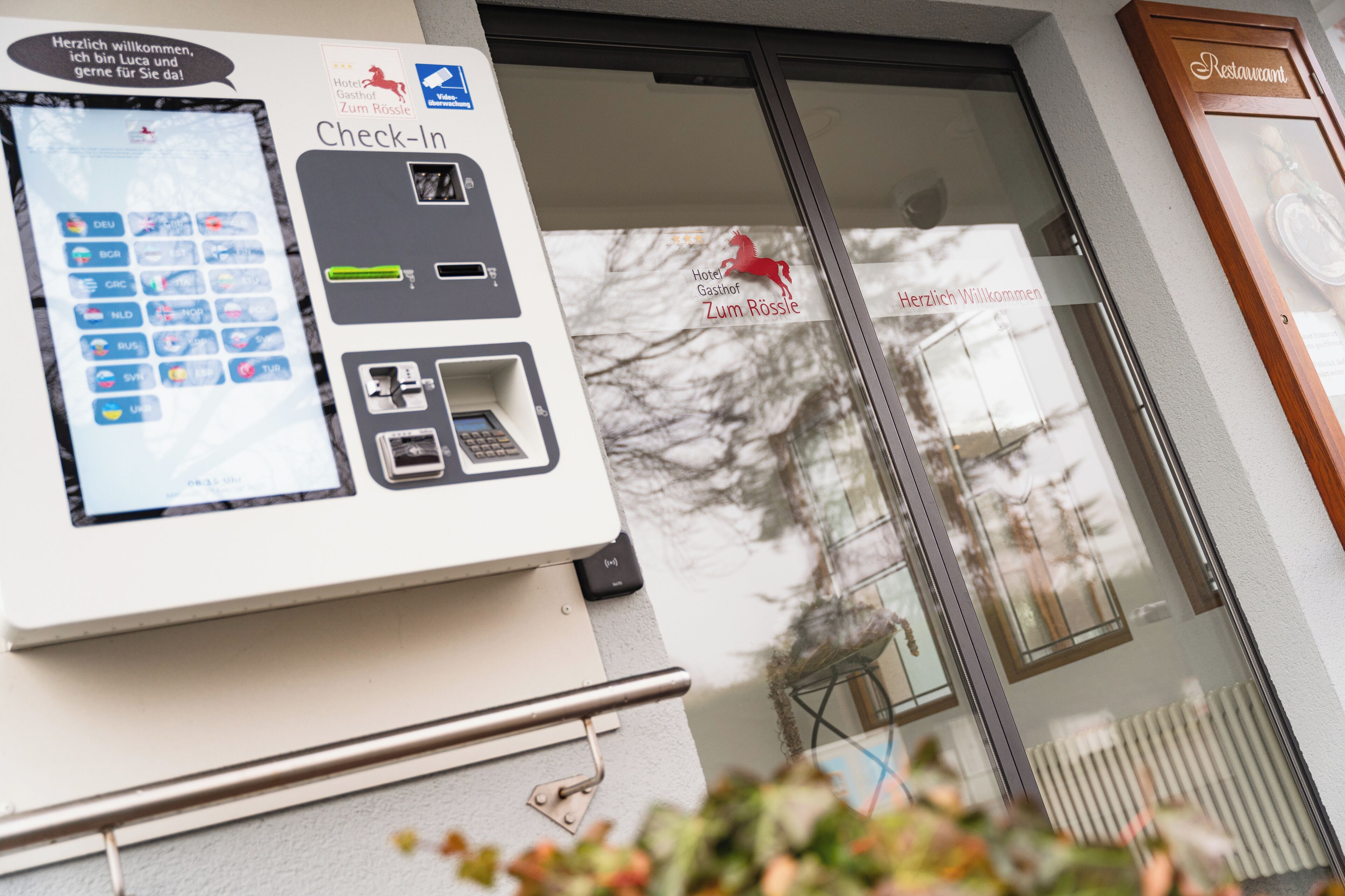 Een incheckautomaat met een digitaal scherm, een bord met een hotelnaam en een bord voor een restaurant.