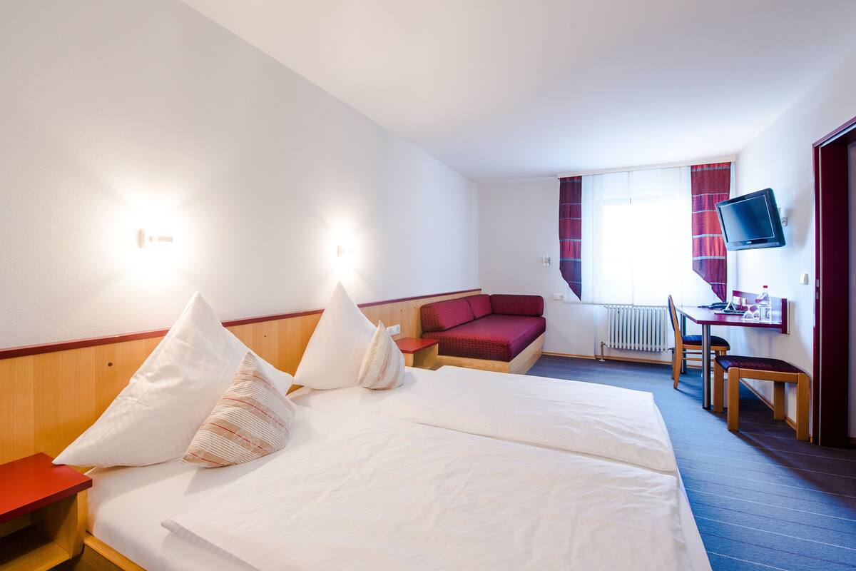 A hotel room with a large bed, a desk, a chair, a TV, and a window with curtains.