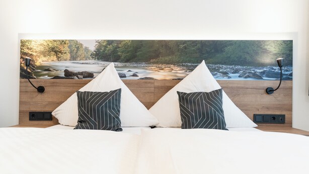 A bed with a view of a river and forest, two pillows, a lamp, and a wall-mounted control panel.