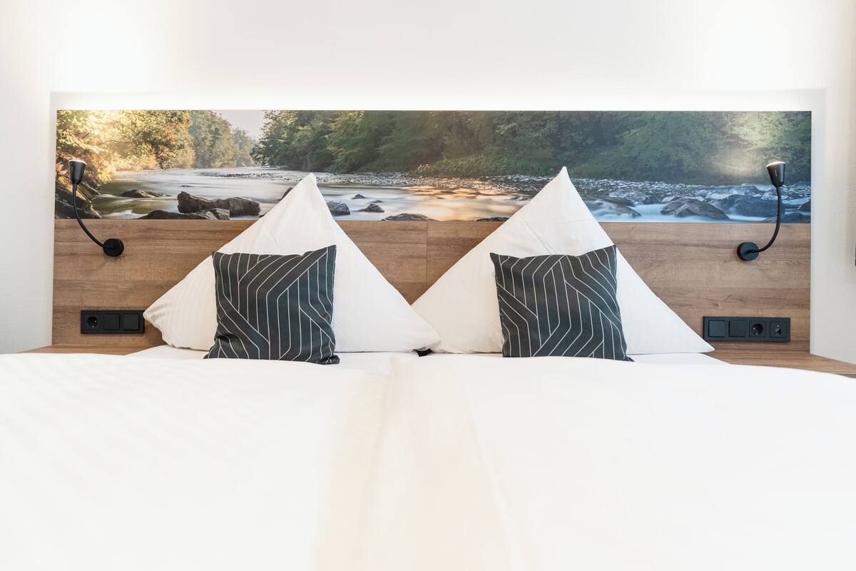 A bed with a view of a river and forest, two pillows, a lamp, and a wall-mounted control panel.