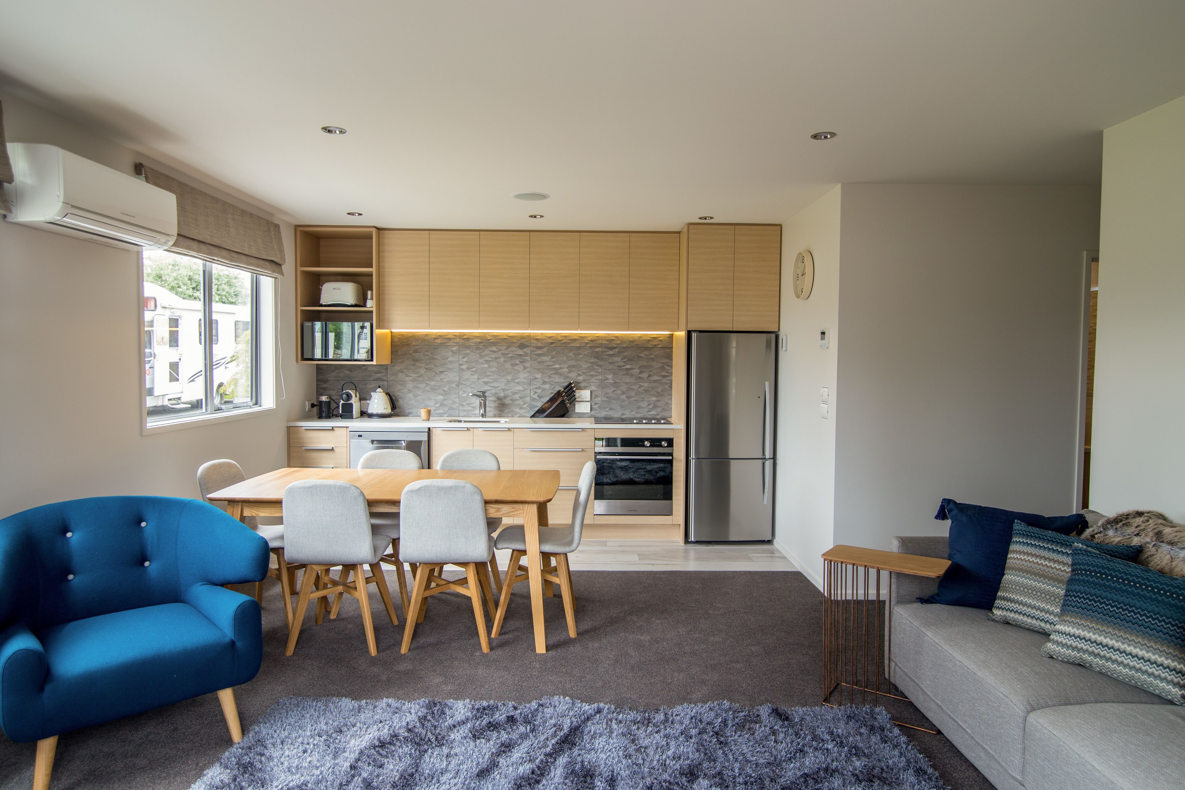 Eine moderne Küche mit Holzausstattung, einem Edelstahlkühlschrank und einem Essbereich mit blauem Stuhl und grauem Sofa.