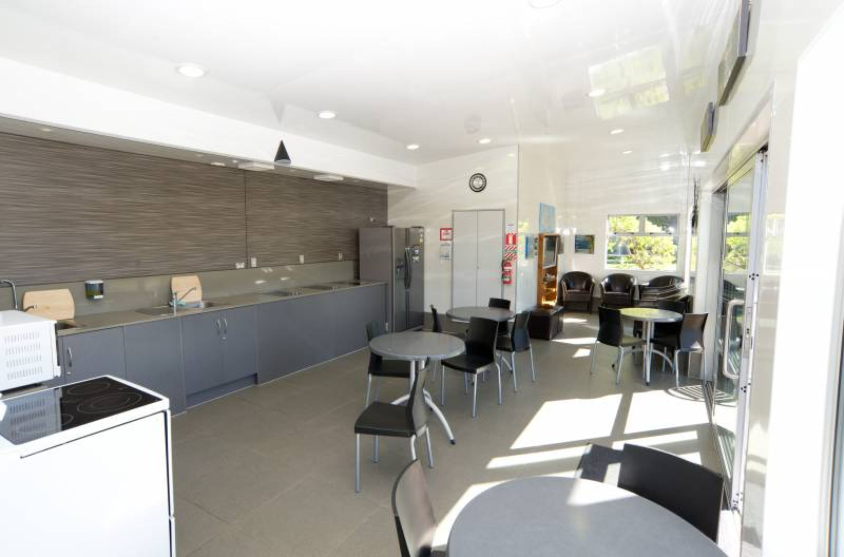 Een moderne keuken met grijze kastjes, een ingebouwde kookplaat en een groot raam. Er staan verschillende tafels en stoelen klaar voor een maaltijd.