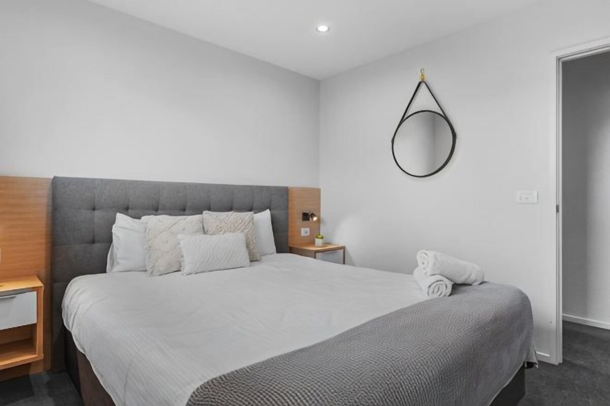 A modern hotel room with a large bed, a bedside table, a wall-mounted mirror, and a door.