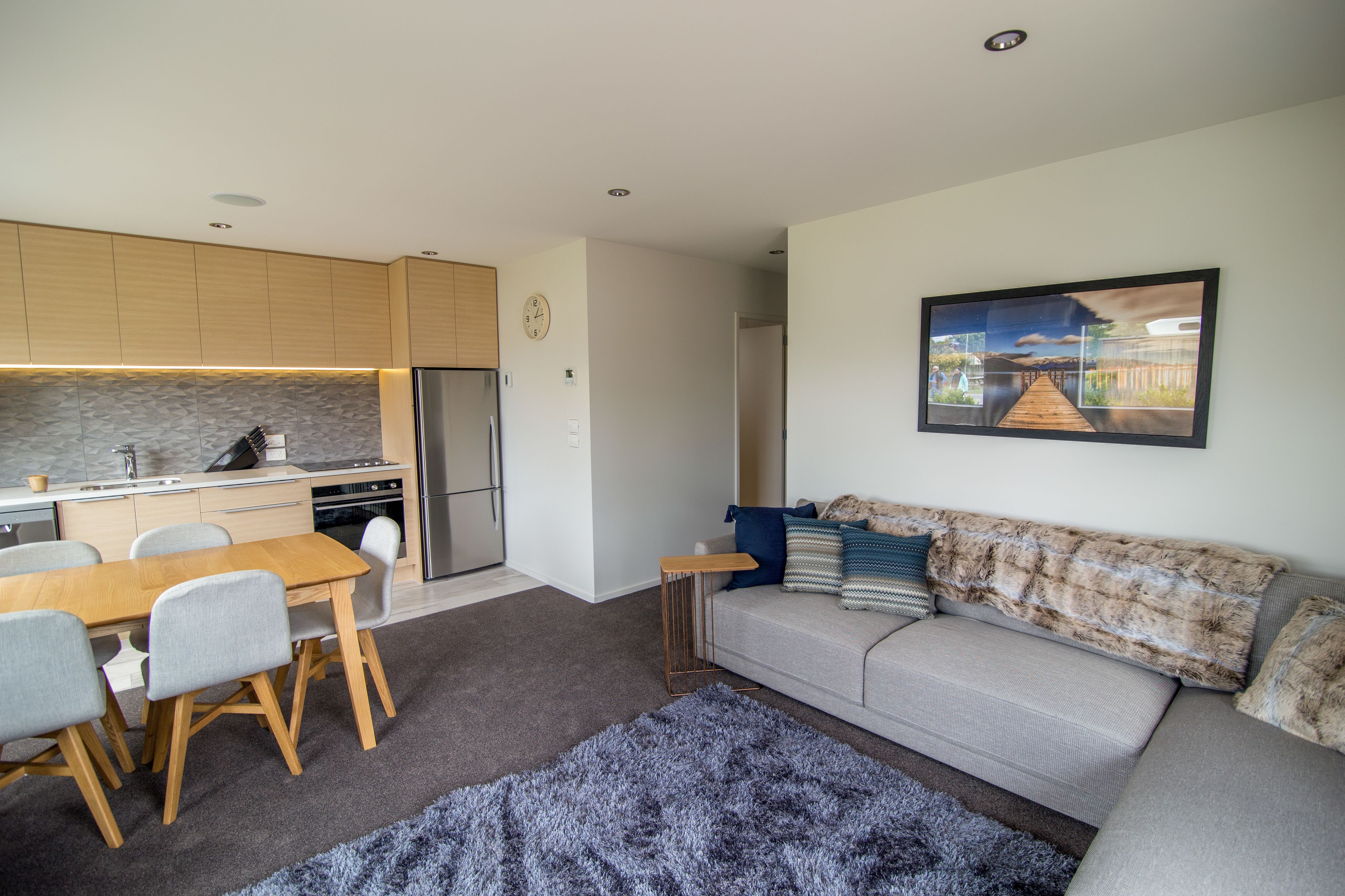 A modern kitchen with wooden cabinets, a dining table with chairs, a grey sofa with patterned cushions, and a large painting on the wall.