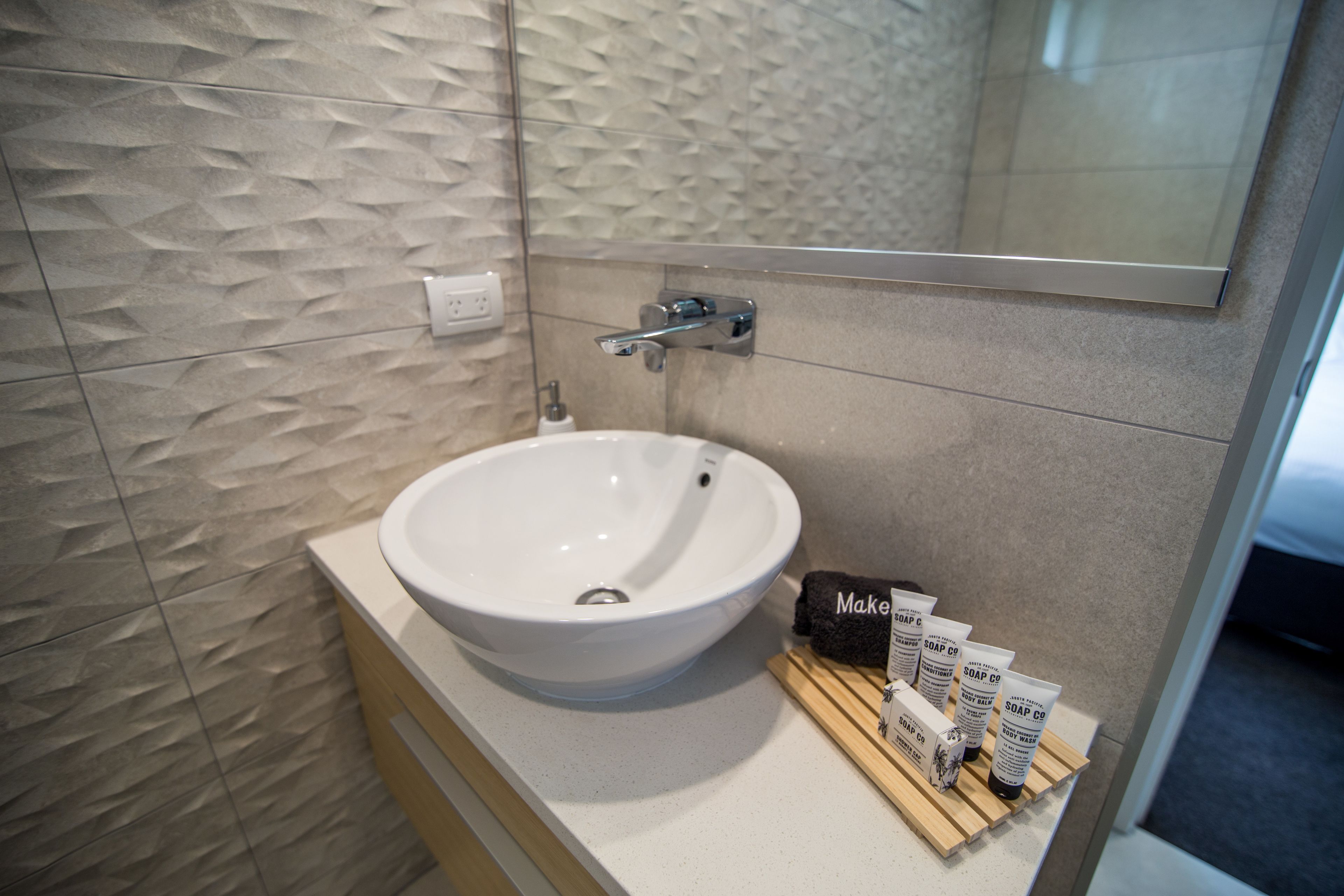A modern bathroom with a white sink, a mirror, and a wooden shelf holding toiletries.