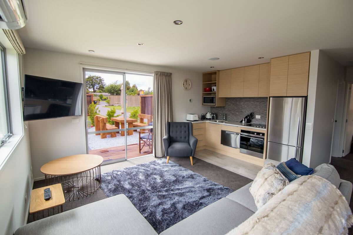 A modern kitchen with a built-in oven and microwave, a grey sofa, a small round table, and a TV mounted on the wall.