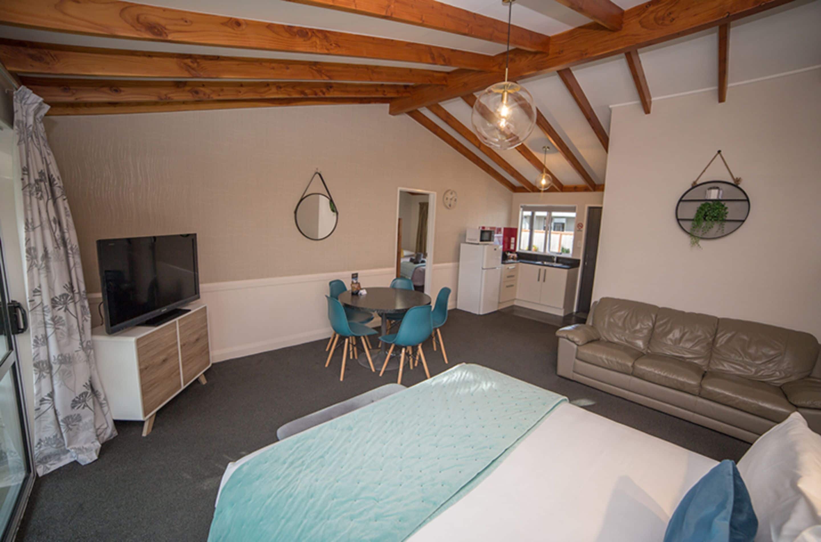 A bedroom with a bed, a TV, a dining table with chairs, a sofa, and a kitchen in the background.
