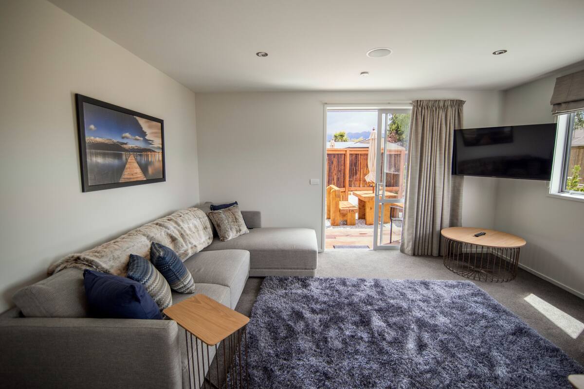 A modern bedroom with a grey sofa, a large bed with a blue blanket, a TV on the wall, and a sliding glass door leading to an outdoor patio.