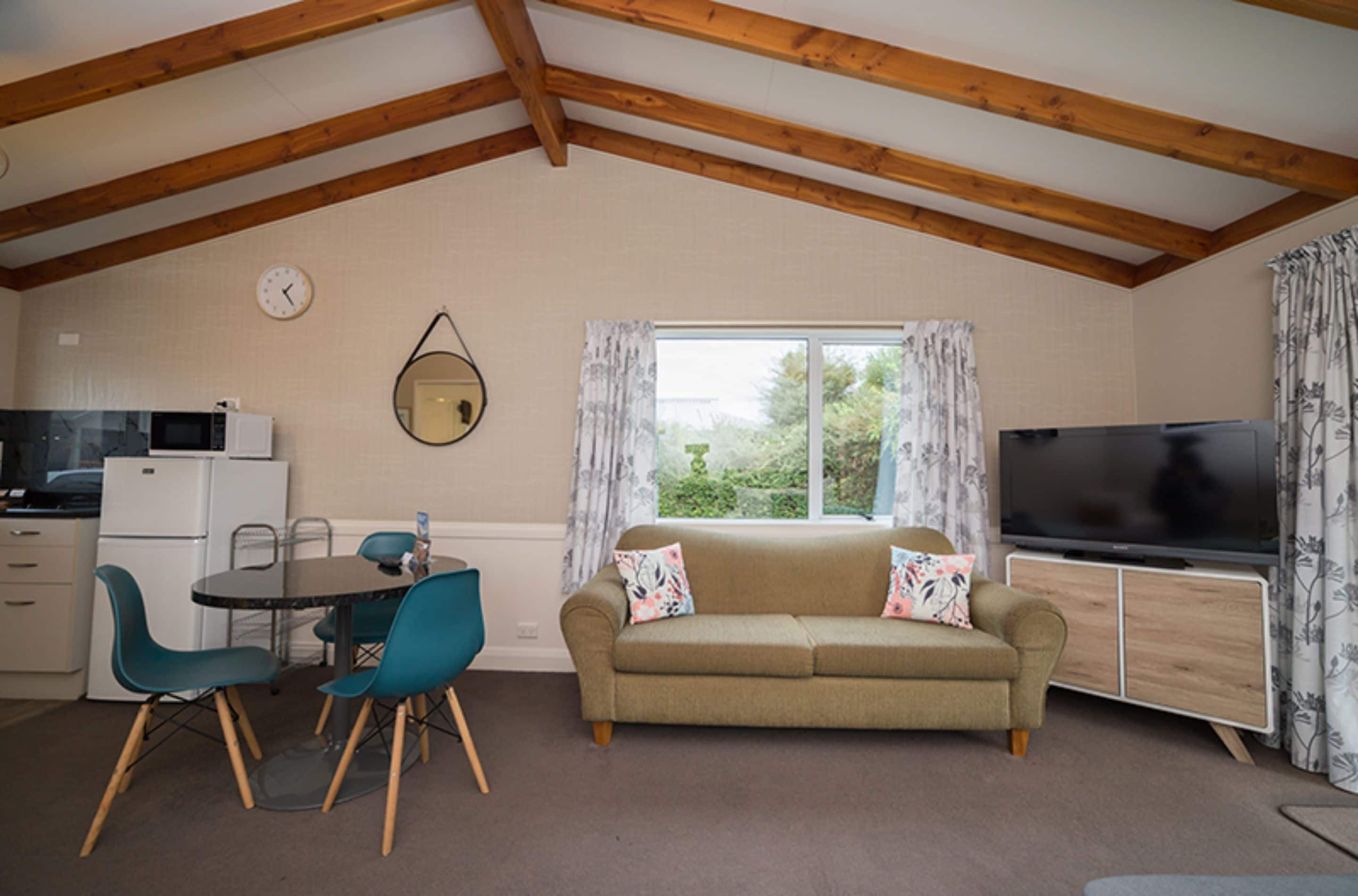 A cozy room with a wooden ceiling, a sofa, a TV, a dining table with chairs, and a kitchen area with a microwave.