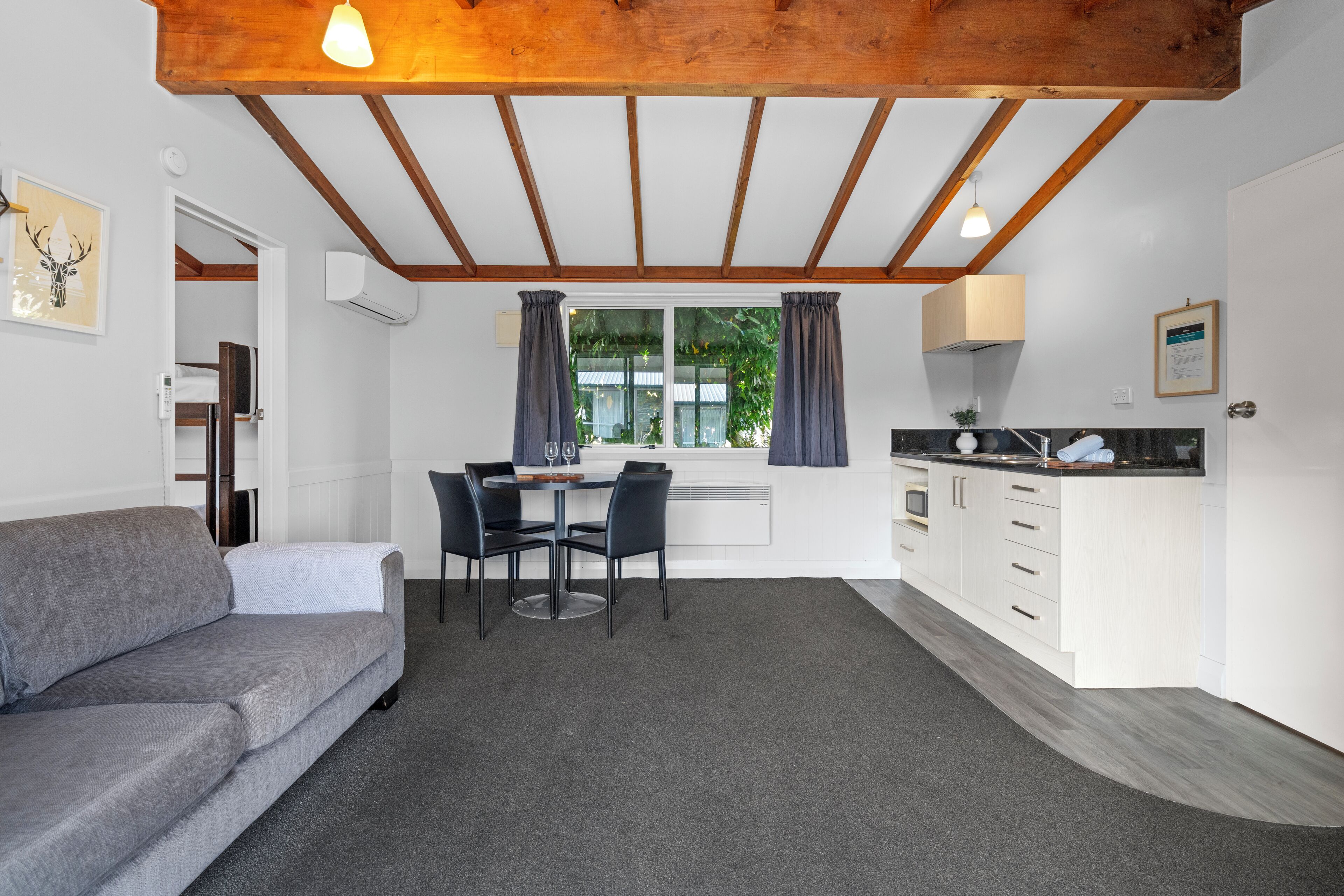 Habitación compacta con un sofá gris, una mesa de comedor con sillas, una kitchenette y una ventana con cortinas.