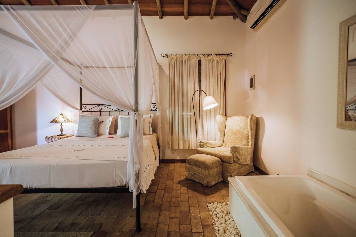 A bedroom with a bed, a chair, a nightstand, a lamp, and a window with curtains.