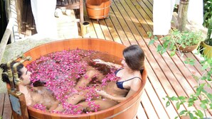 Una terraza de madera con una bañera grande de madera llena de agua y flores rosadas, rodeada de plantas en macetas y una mesa blanca con sillas.