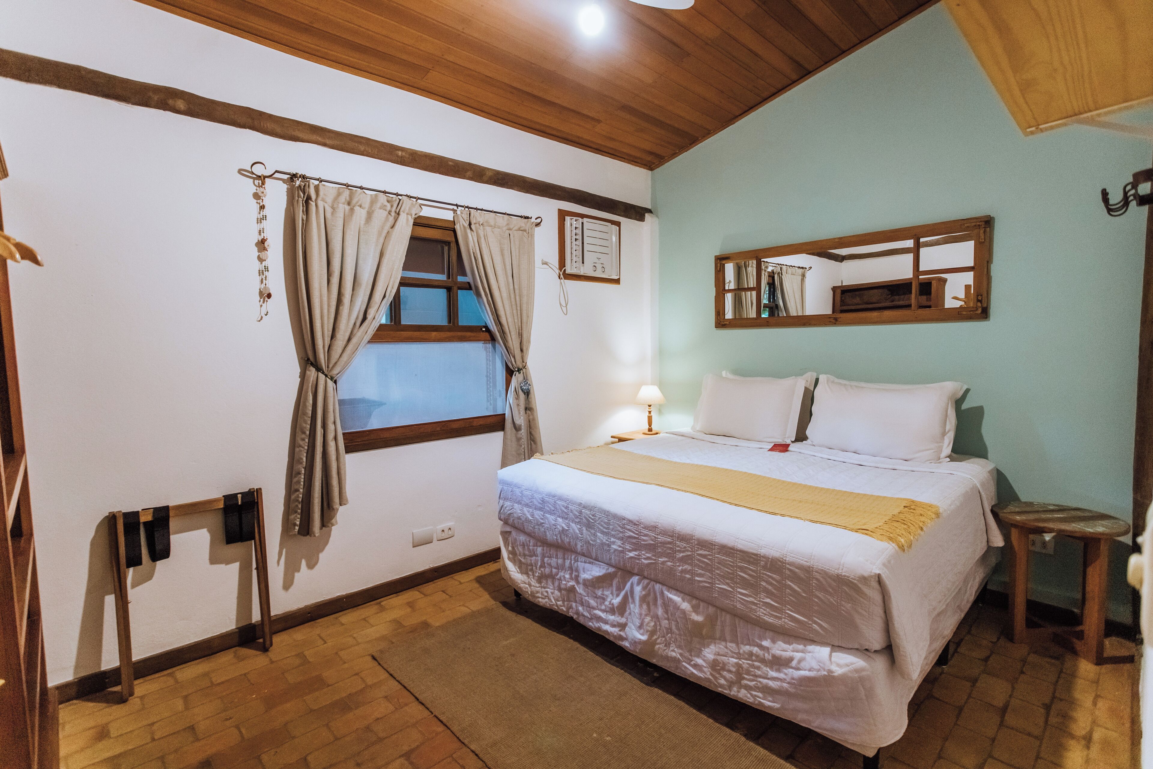 Un dormitorio con una cama, una ventana con cortinas, un espejo y un taburete pequeño.