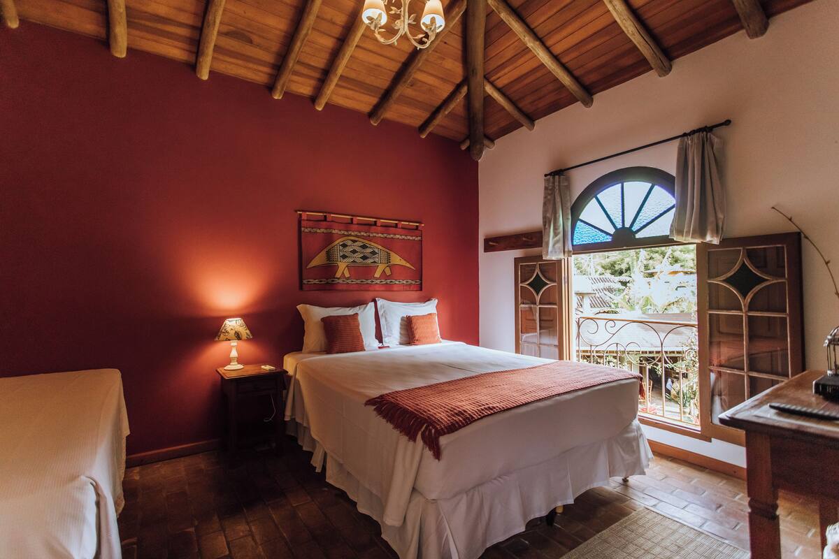 A bedroom with a wooden ceiling, a bed with white bedding, a nightstand with a lamp, and a window with a view of greenery.