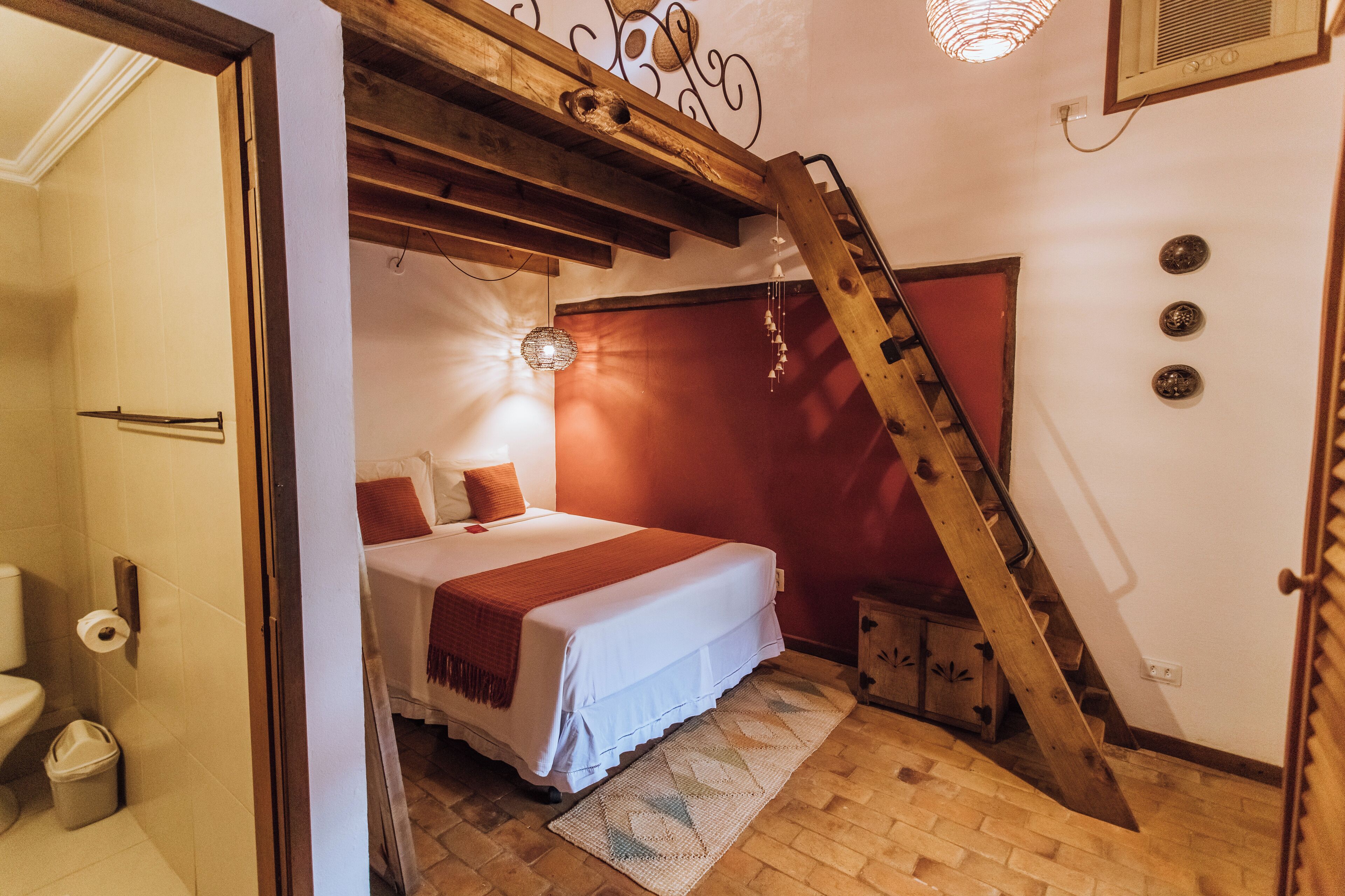 A bedroom with a bed, wooden beams, a red wall, a staircase, and a small cabinet.