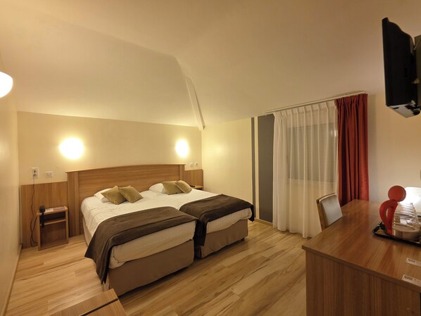 A hotel room with two beds, a wooden headboard, bedside tables, a desk with a chair, a TV, and a window with curtains.