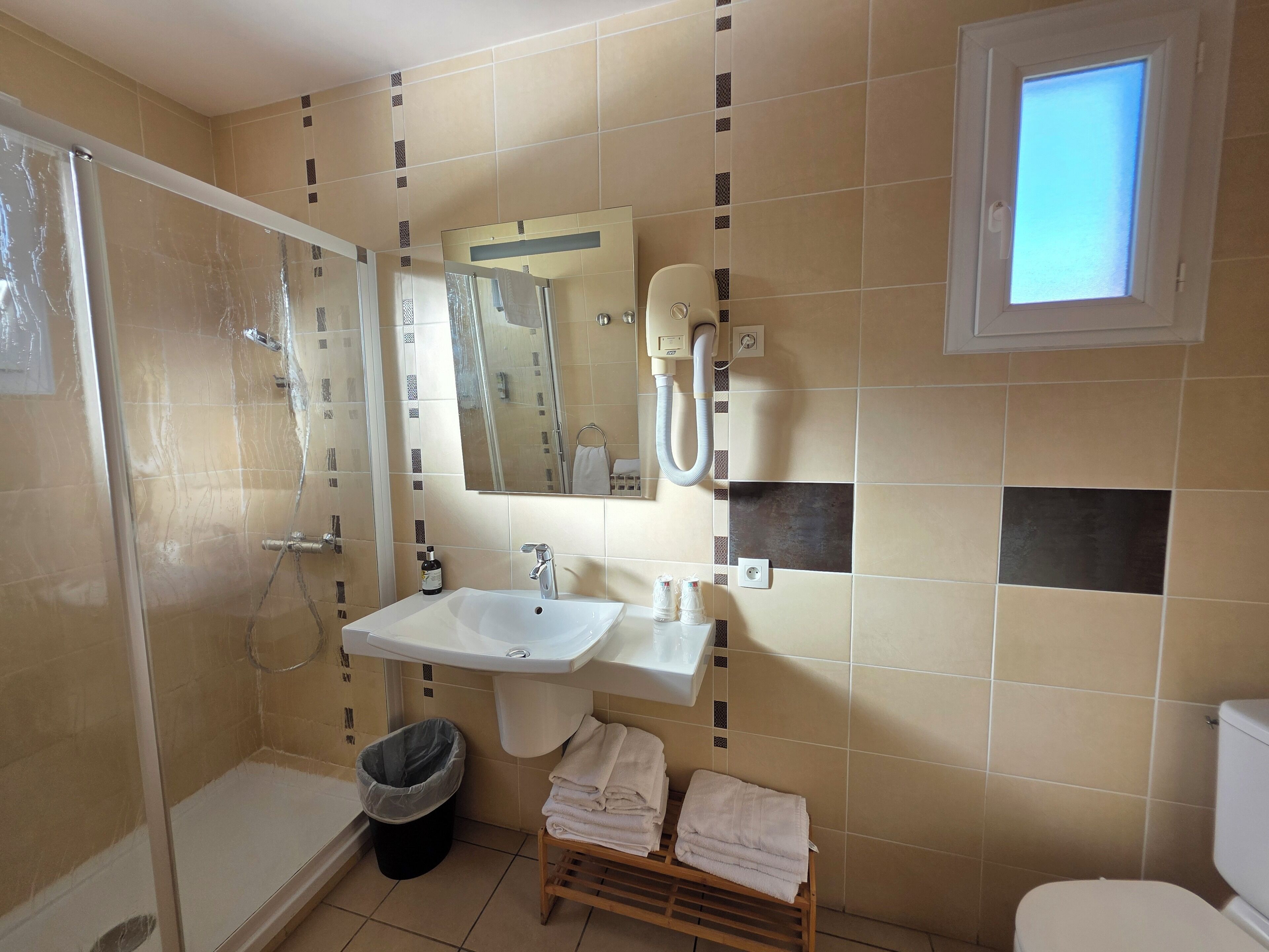 Een badkamer met een douche, wastafel en toilet. Boven de wastafel hangt een spiegel, er is een raam en een handdoekrek met handdoeken.