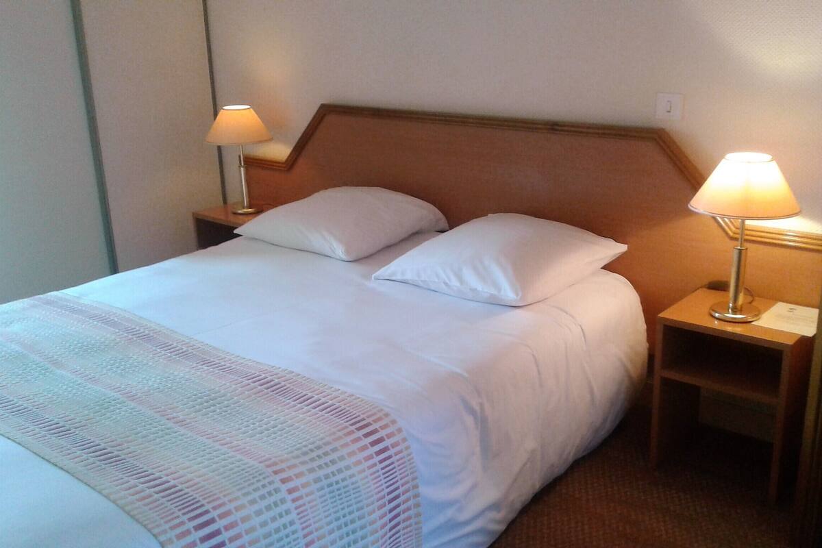 A neatly made bed with white linens and two pillows, flanked by two bedside tables with lamps, in a room with a simple wooden headboard and plain walls.