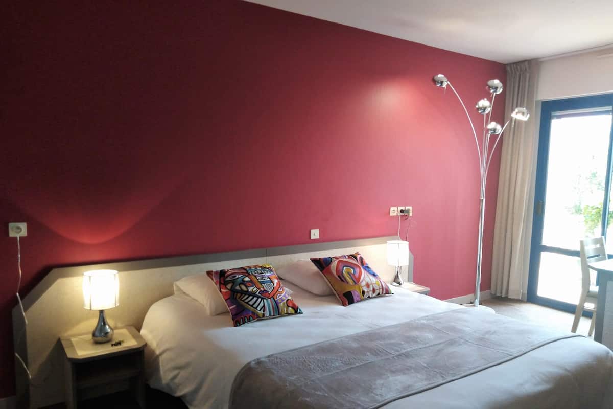 A hotel room with a large bed, two bedside tables with lamps, a modern floor lamp, and a view of the outdoors through a large window.