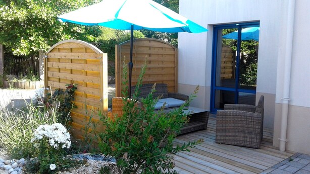 Un patio con valla de madera, una sombrilla azul, mobiliario de mimbre y un camino de piedra.