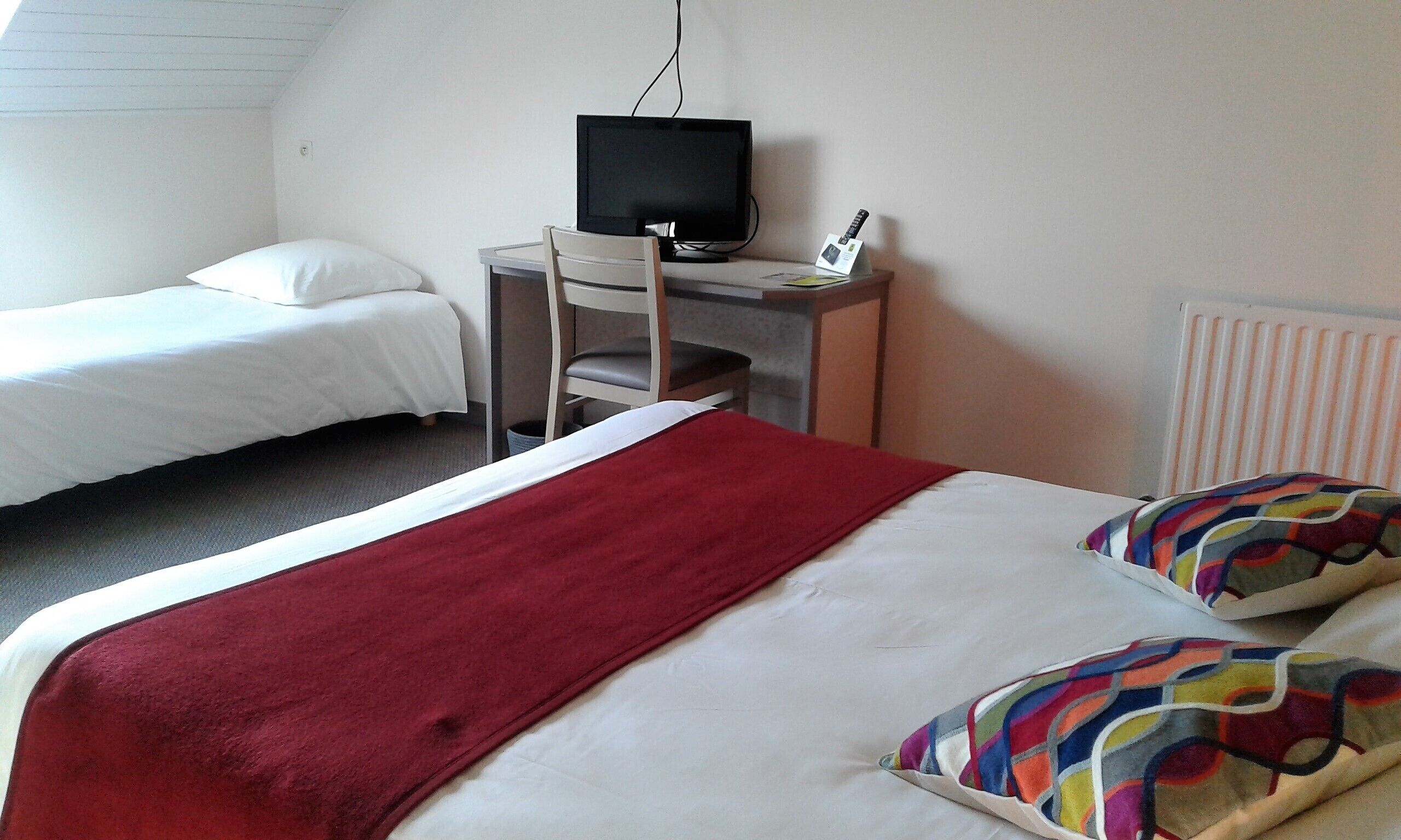A hotel room with two single beds, a red blanket on one bed, a television on a small table, and a radiator on the wall.