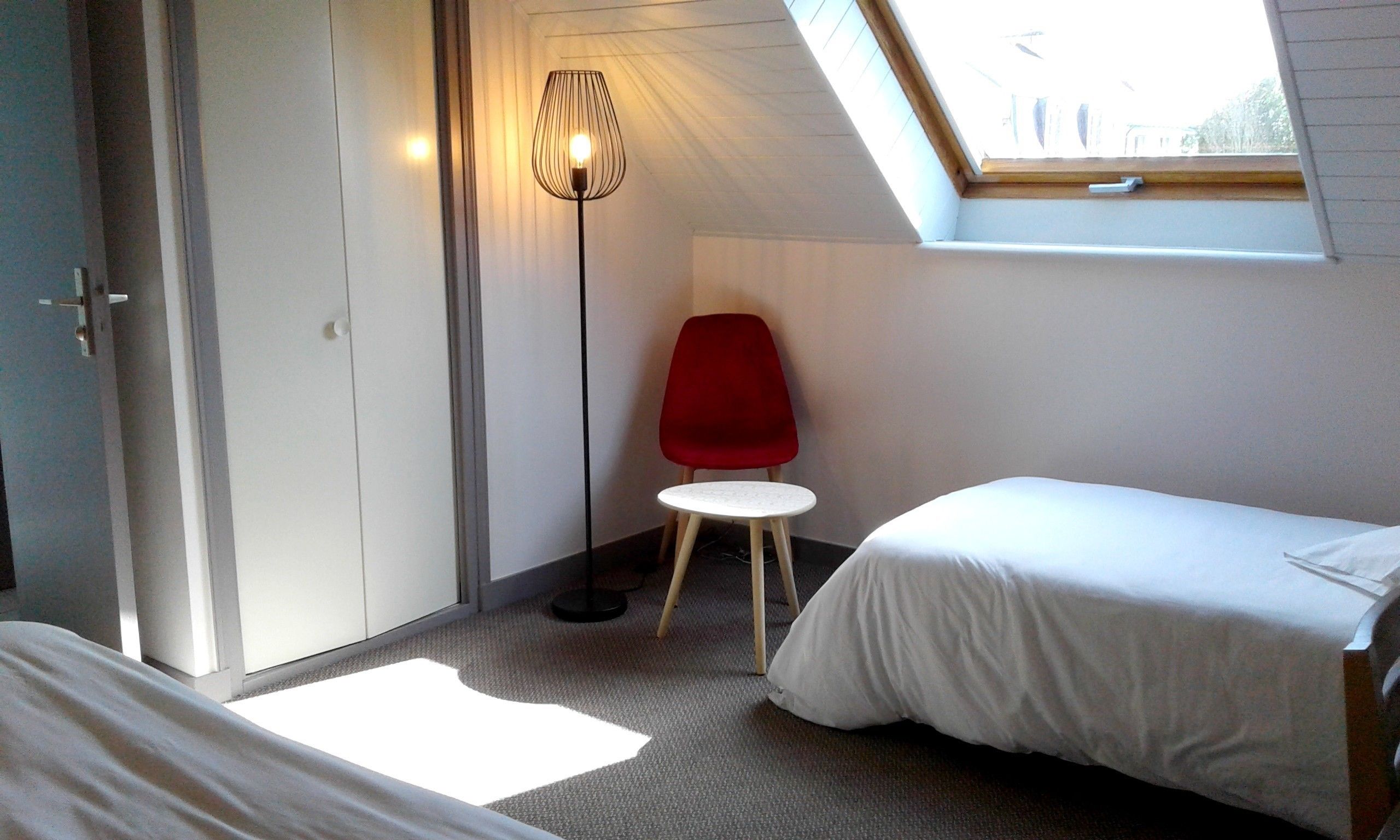Ett litet, välordnat sovrum med en enkel säng, en röd stol och en golvlampa.