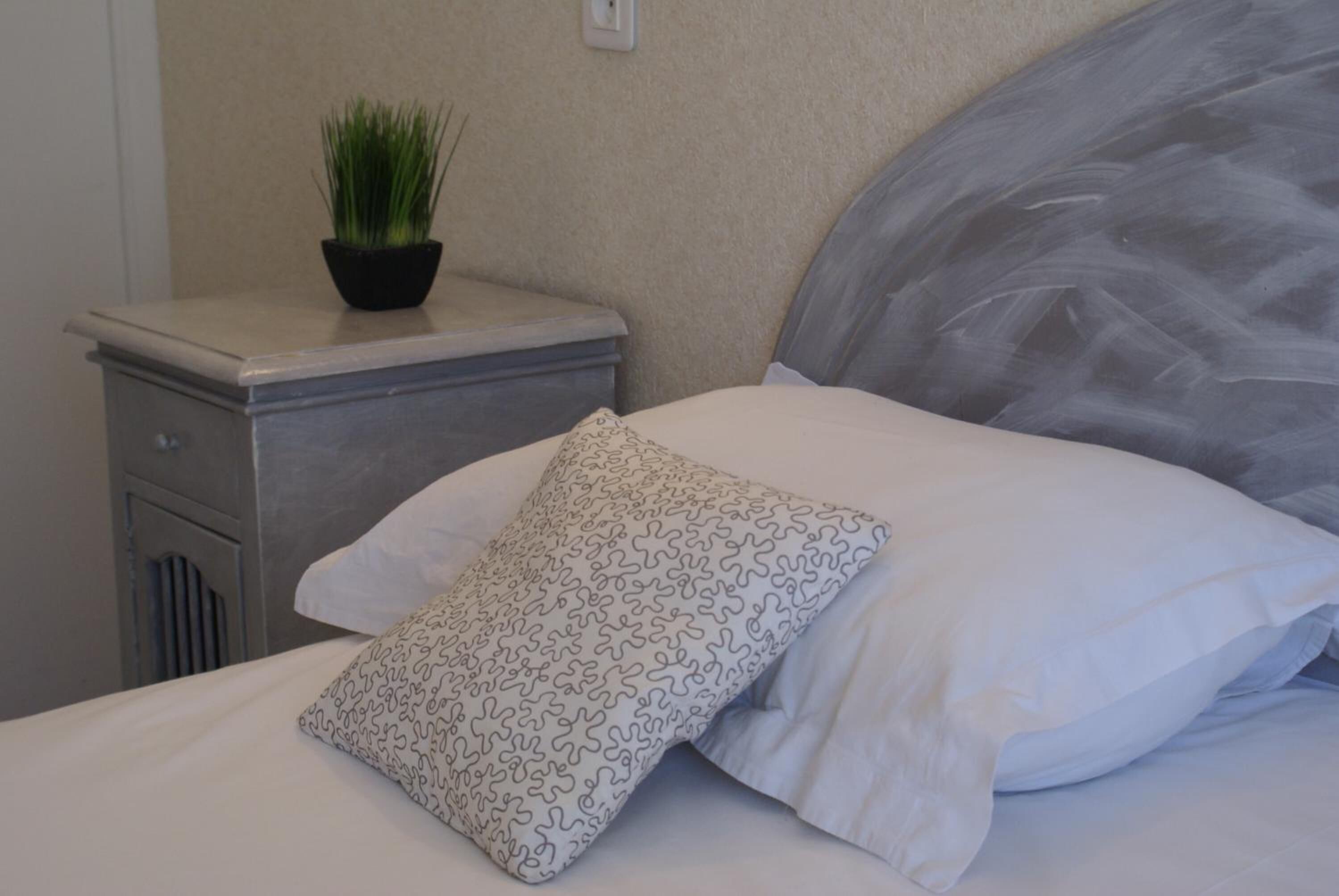 A neatly made bed with a white pillow and a patterned pillow, a small potted plant on a bedside table, and a wall with a textured finish.
