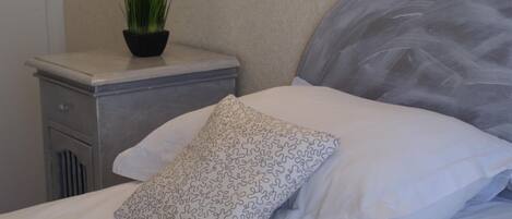 A neatly made bed with a white pillow and a patterned pillow, a small potted plant on a bedside table, and a wall with a textured finish.