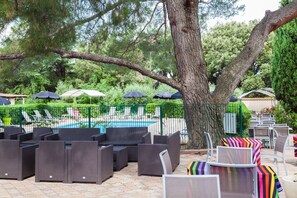 An outdoor seating area with wicker furniture and umbrellas, a pool, and a tree. - Hôtel Le Petit Manoir Provence (Les Angles)