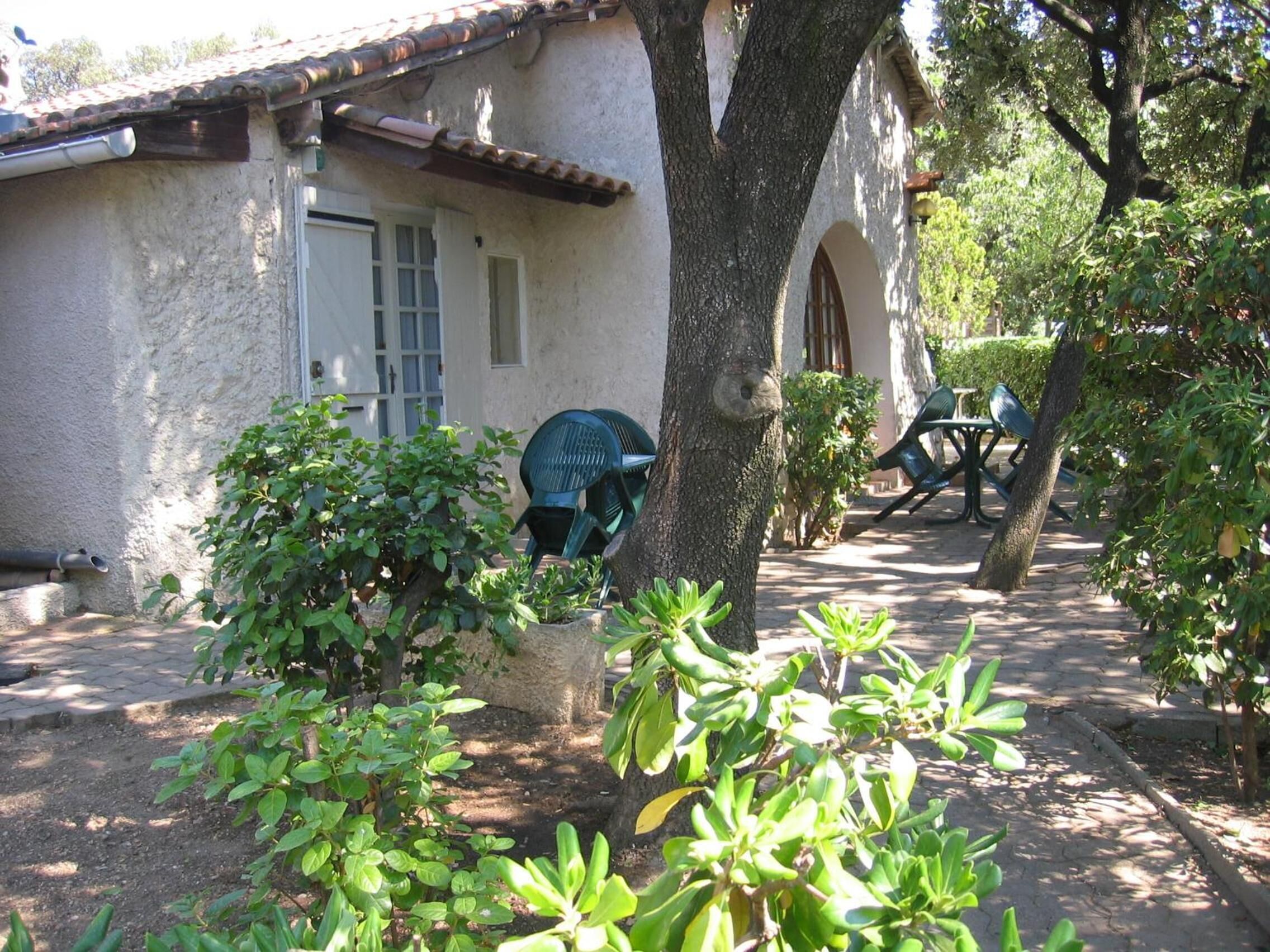 A rustic building with a tiled roof, a door, and windows. There are green plants and a tree in the foreground.