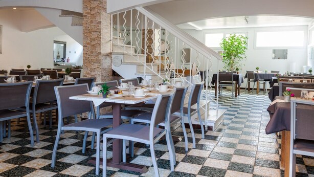 A spacious dining area with checkered flooring, multiple tables and chairs, a stone pillar, and a staircase with a decorative railing. - Hôtel Le Petit Manoir Provence (Les Angles)