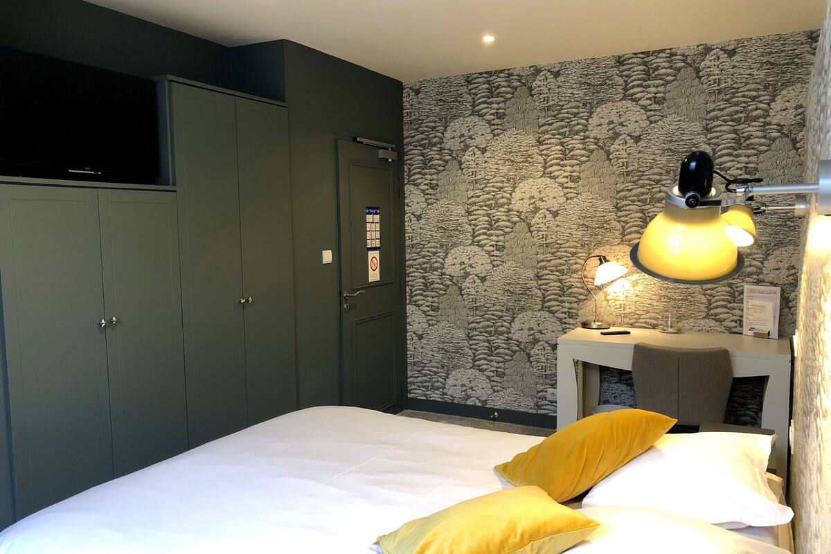 A hotel room with a bed, a desk with a lamp, a wardrobe, and a patterned wall.