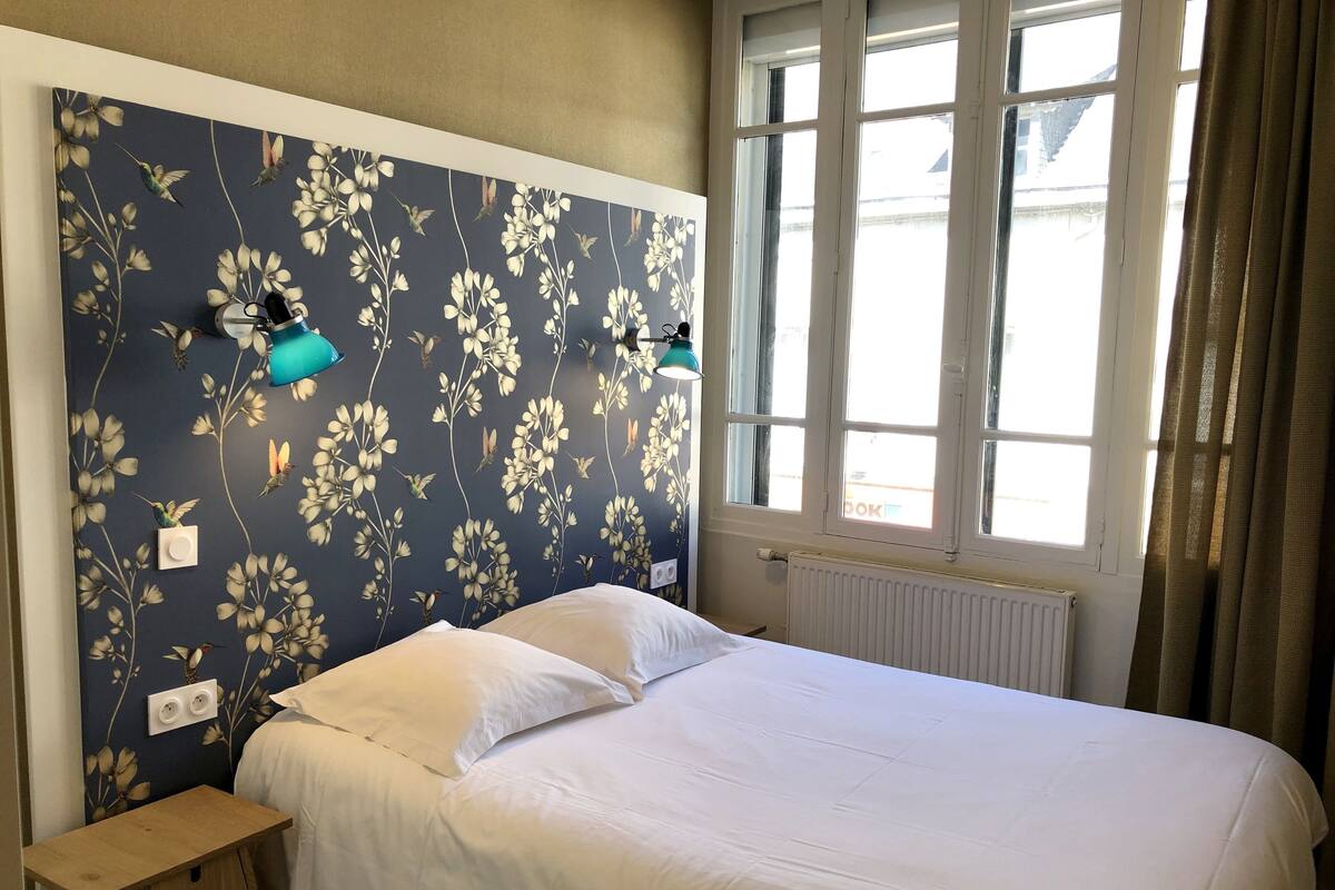 A bedroom with a floral wallpaper, a bed with white linens, a bedside table, and a window with curtains.