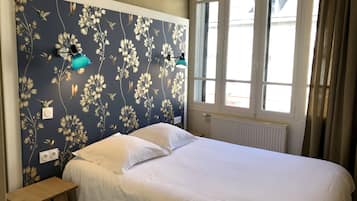 A bedroom with a floral wallpaper, a bed with white linens, a bedside table, and a window with curtains.