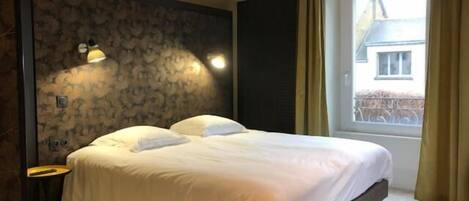 A neatly made bed in a room with a patterned wall, a small table, and a window with curtains.