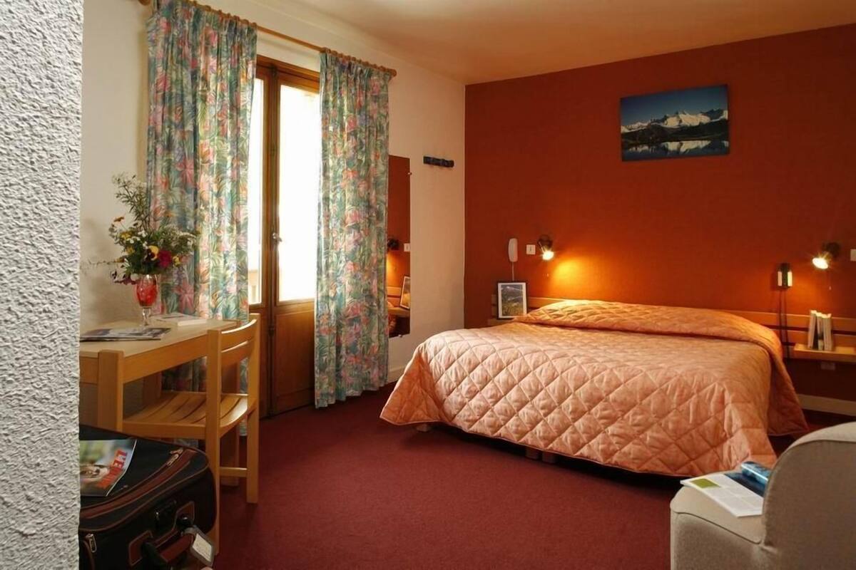 A hotel room with a bed, a desk, a chair, a suitcase, and a window with curtains.