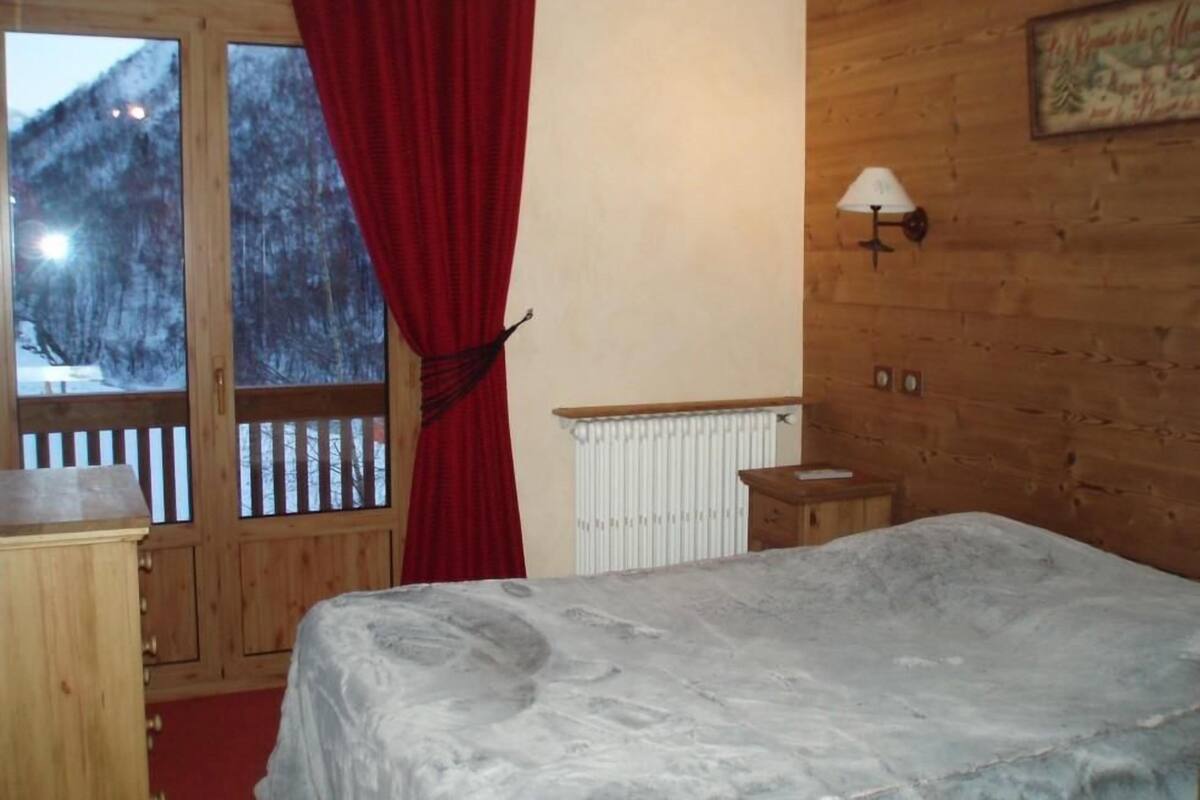 A bedroom with a bed, a window with a red curtain, a wooden wall, and a small bedside table.