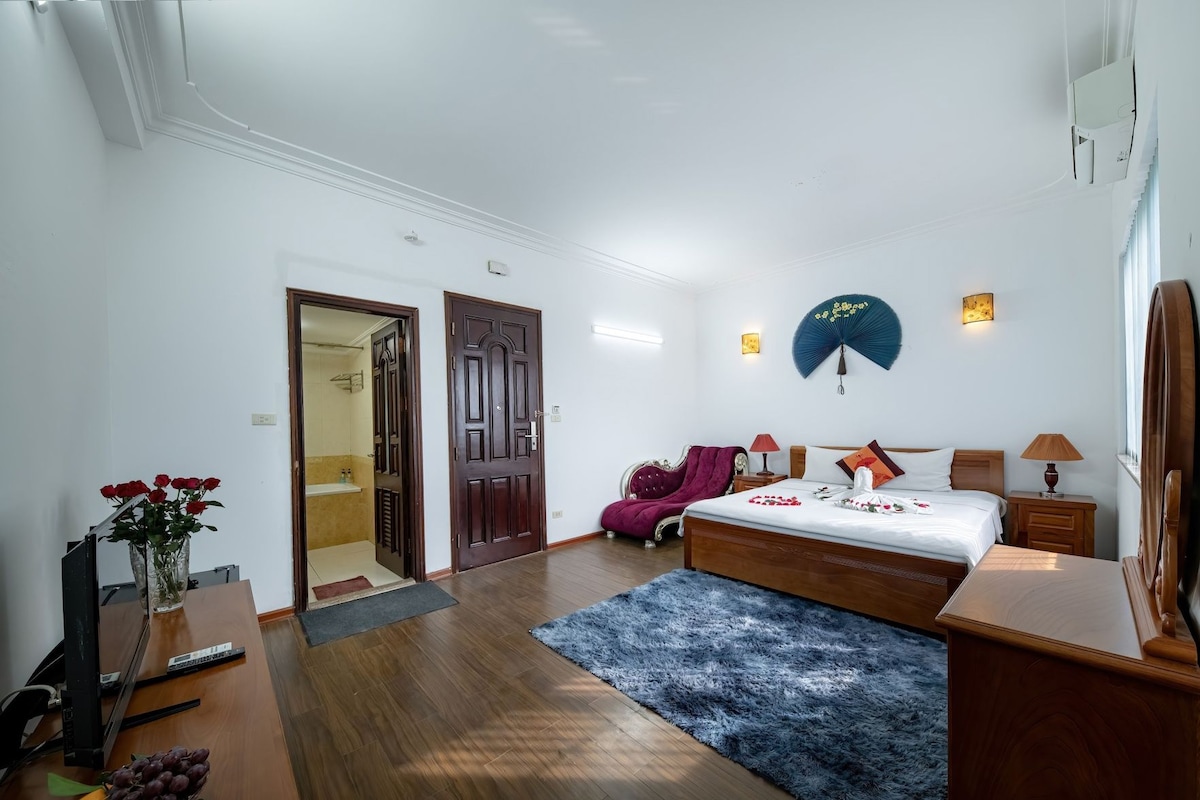 Una habitación de hotel con una cama, mesitas de noche, tocador, espejo, una silla roja, alfombra azul y un jarrón con flores.