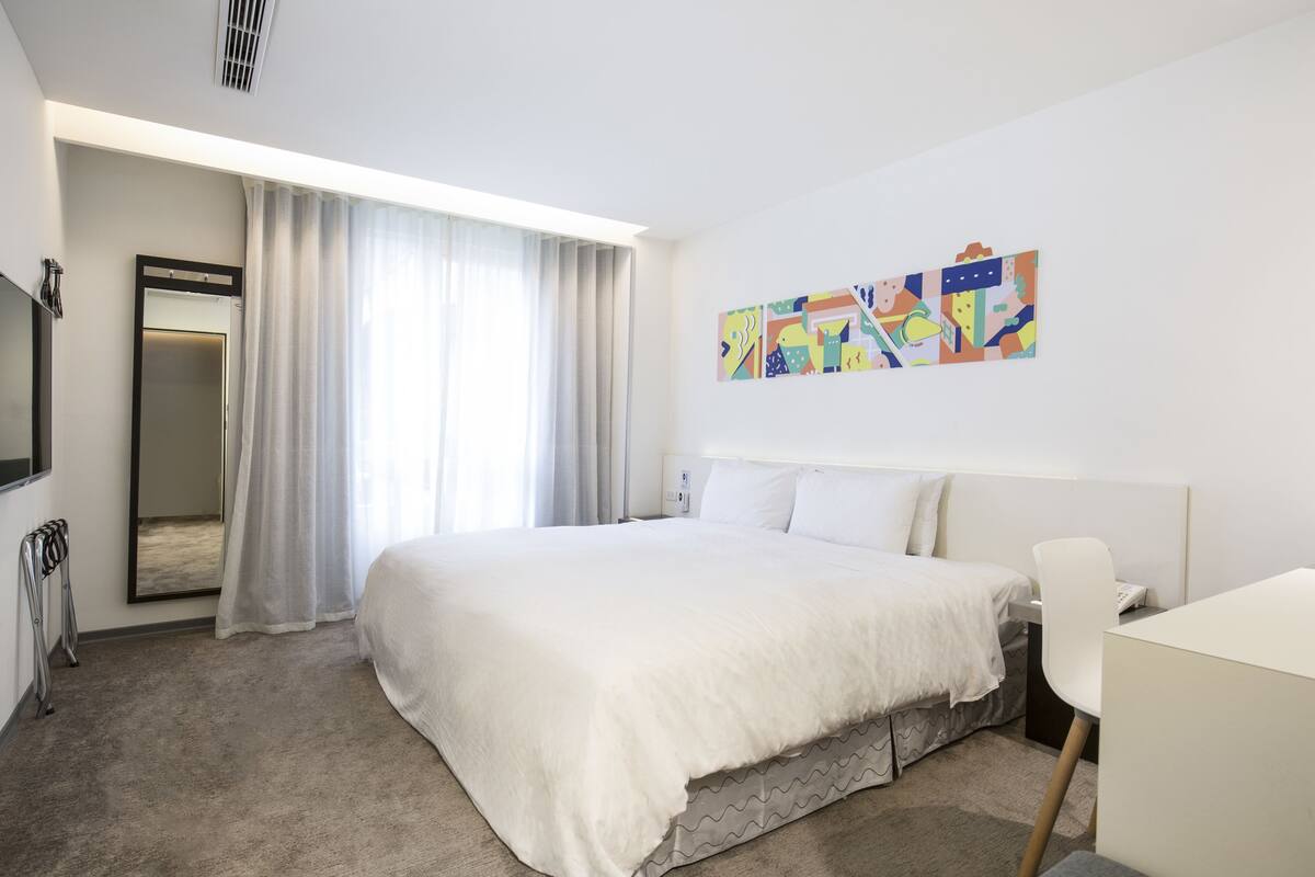Una habitació d'hotel amb un llit gran, un escriptori, una cadira, un mirall i un quadre abstracte i colorit a la paret.