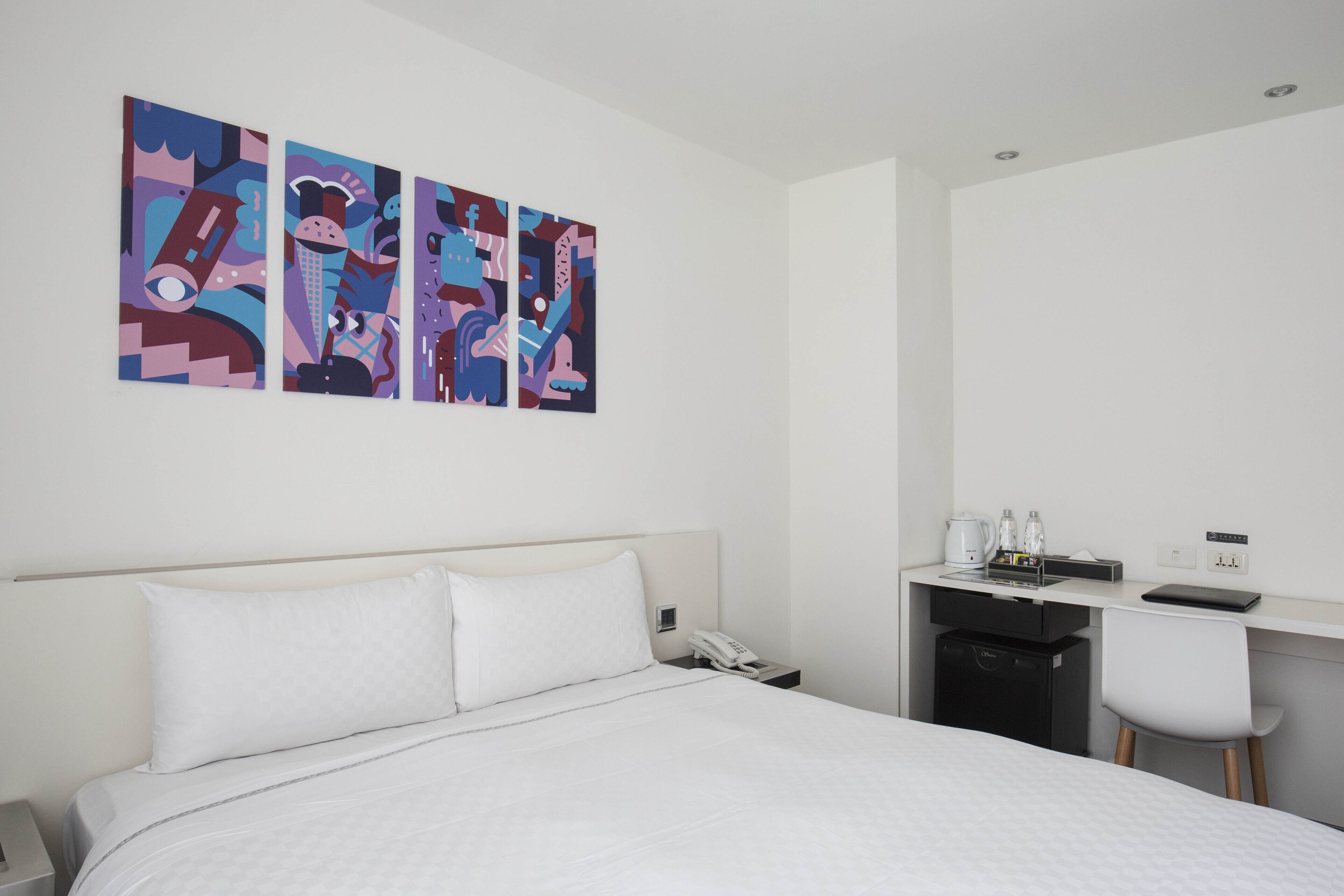 A hotel room with a bed, a desk, and a chair. There are three paintings on the wall.