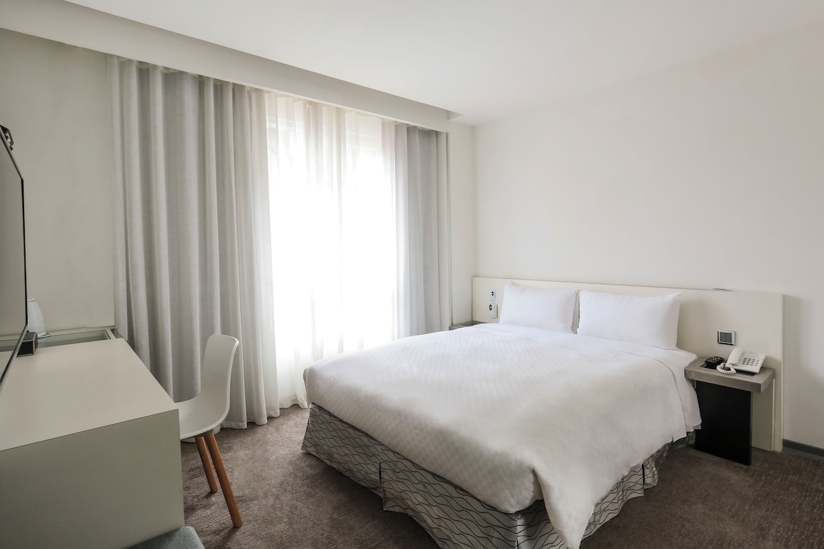 Une chambre d’hôtel avec un lit, un bureau, une chaise, une table de chevet et une fenêtre avec des rideaux.