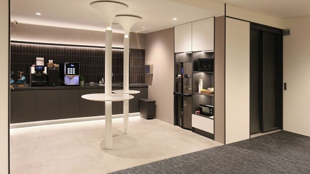 Un hall d’hôtel moderne comprenant un comptoir d’accueil, un kiosque en libre-service, un espace détente avec une table blanche et un ascenseur à la structure noire.