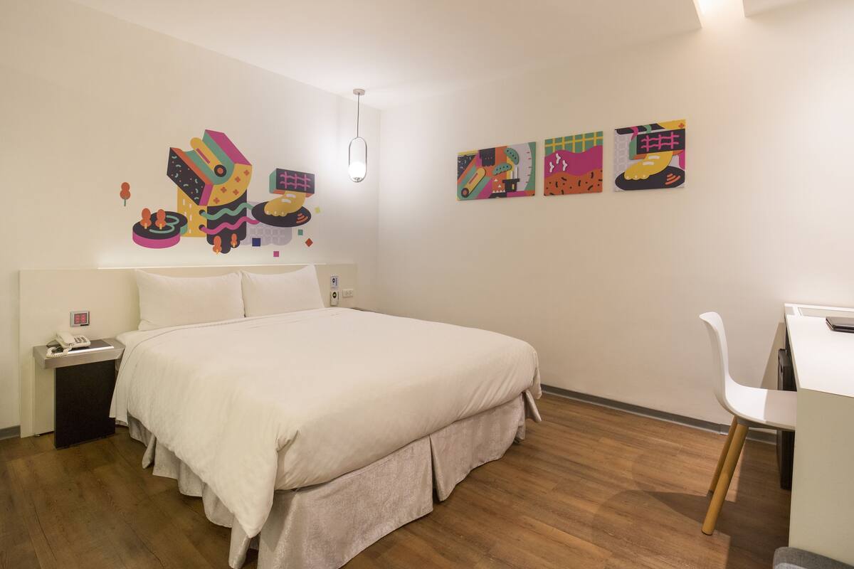 Una habitació d'hotel amb un llit gran, un escriptori amb una cadira i obra d'art colorida a les parets.