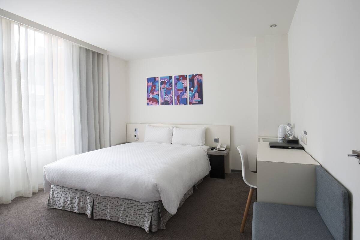 Una habitació d'hotel moderna amb un llit gran, un escriptori i una cadira. Hi ha tres quadres a la paret i una finestra amb cortines.