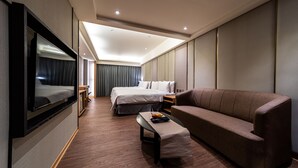 A modern hotel room with a large bed, a sofa, a TV mounted on the wall, and a small table with fruits.