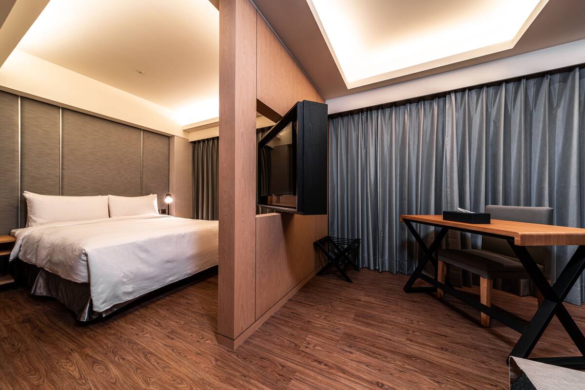 Una camera d'albergo moderna con un letto, una scrivania, una sedia, una TV e un'ampia finestra con tende.
