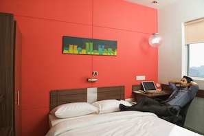 Quarto de hotel com parede vermelha, cama com roupa de cama branca, escrivaninha com um laptop e uma pintura de paisagem urbana.